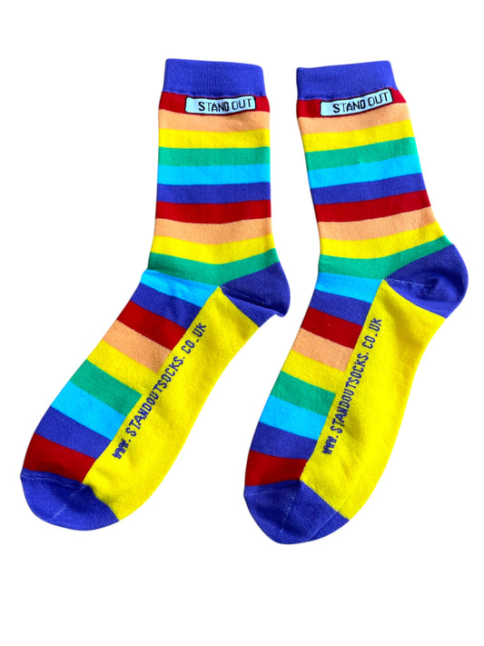 A photograph of a pair of Stand Out Socks Rainbow Pride design, showcasing vibrant rainbow stripes and the Stand Out logo near the cuff and website on the foot.