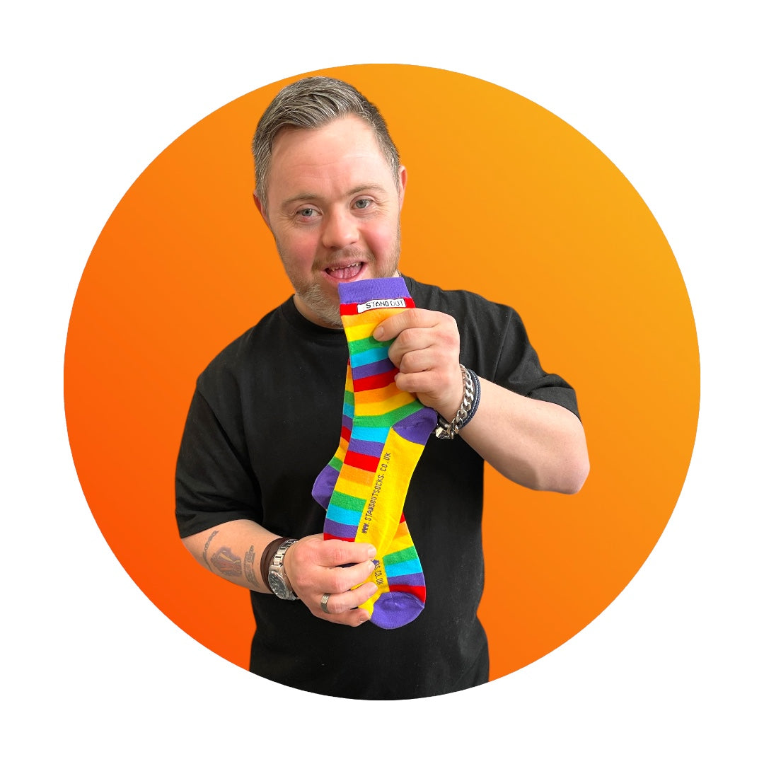 A smiling man with Down syndrome proudly holding up the Stand Out Socks Rainbow Pride socks, framed by a bold orange circle in the background.