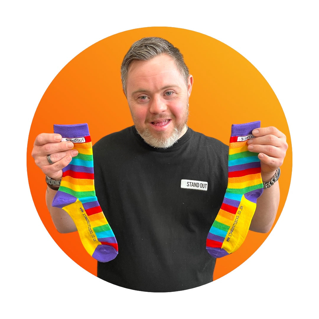 A smiling man with Down syndrome proudly holding up the Stand Out Socks Rainbow Pride socks, framed by a bold orange circle in the background.