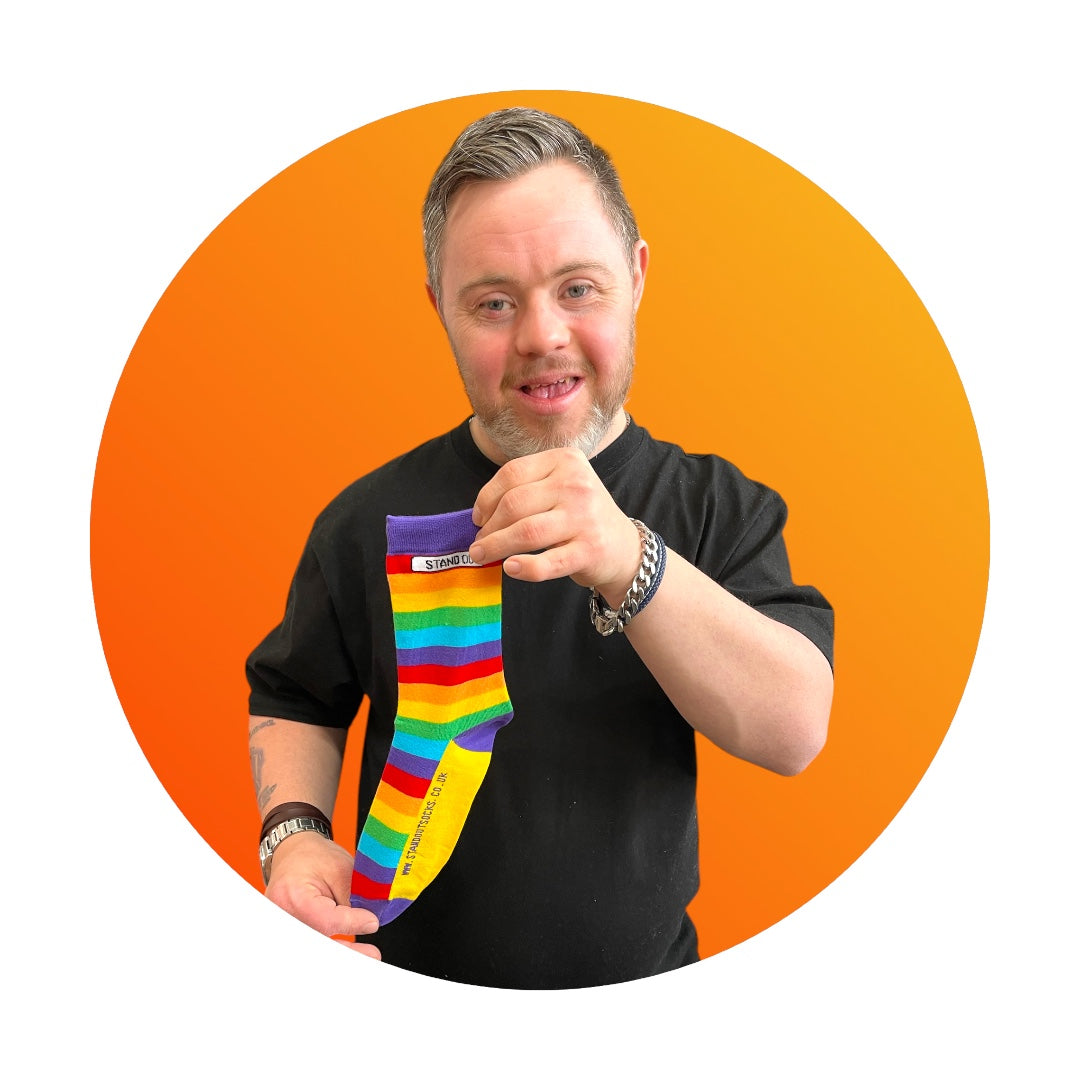 A smiling man with Down syndrome proudly holding up the Stand Out Socks Rainbow Pride socks, framed by a bold orange circle in the background.