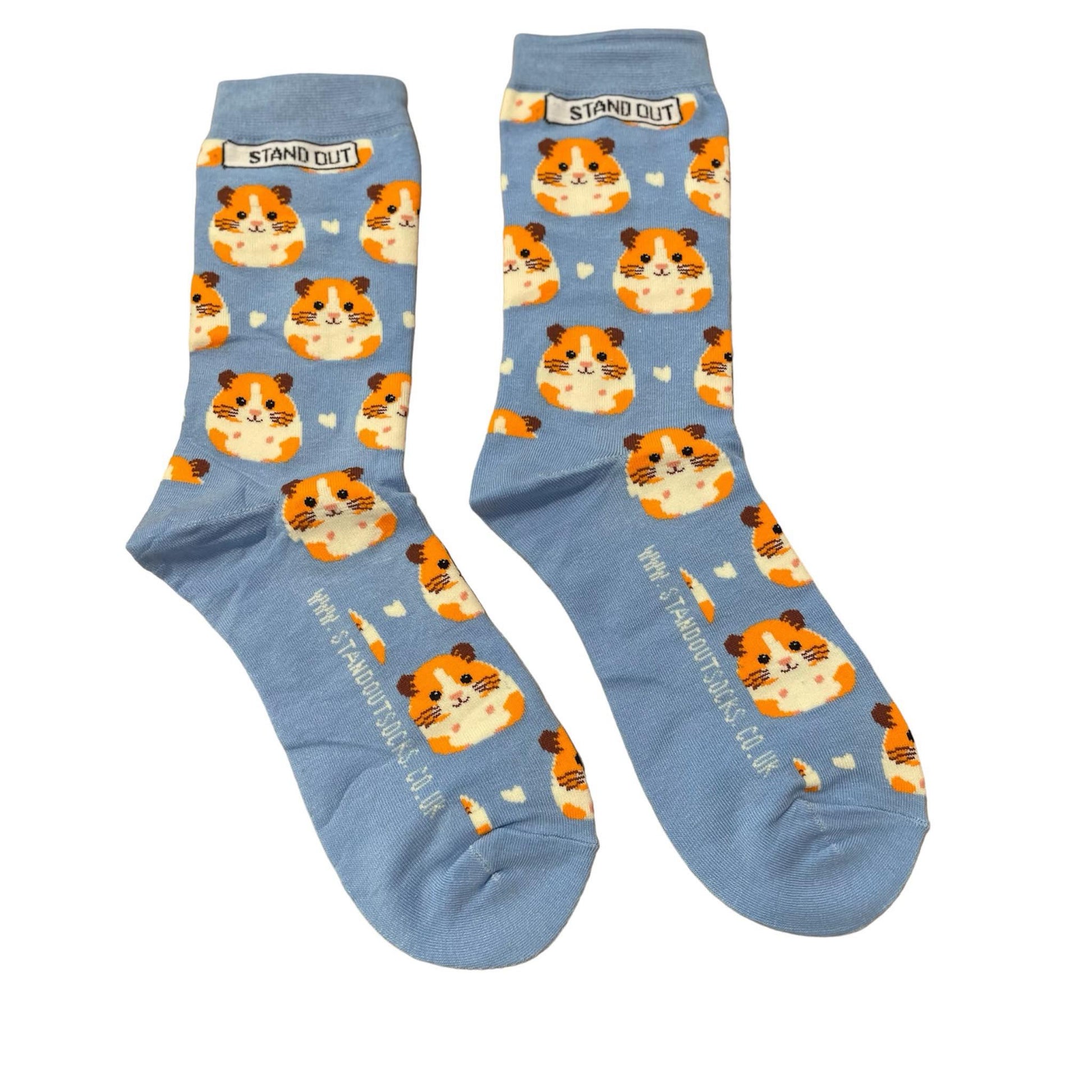 Light blue socks featuring a cute guinea pig pattern, created by Stand Out Socks. The socks display a playful design with orange and white hamster faces and small white hearts scattered throughout. The words "STAND OUT" are visible on the cuff and the company's website is printed on the sole.
