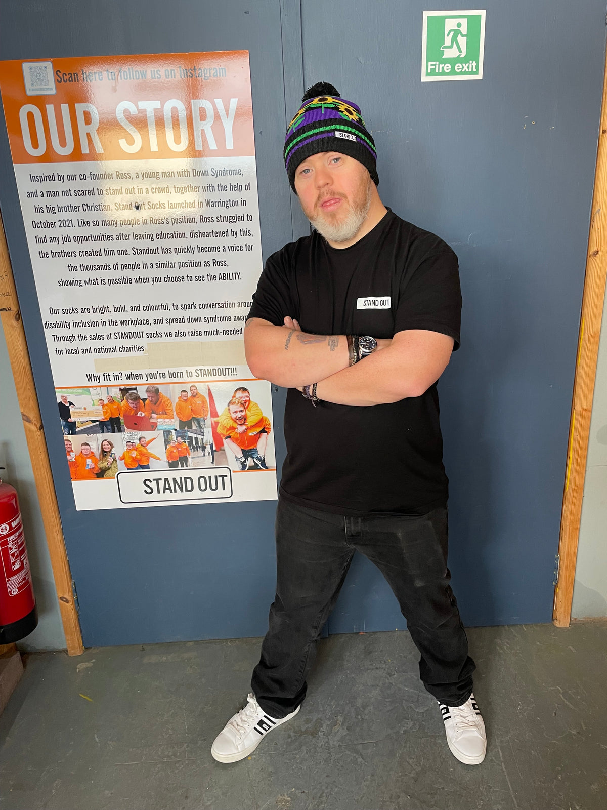 A man wearing a black t-shirt, dark jeans, and a colorful striped beanie stands with arms crossed in front of a blue door. To his left is a large printed 'Our Story' poster with images of soccer players and the text 'STAND OUT'.