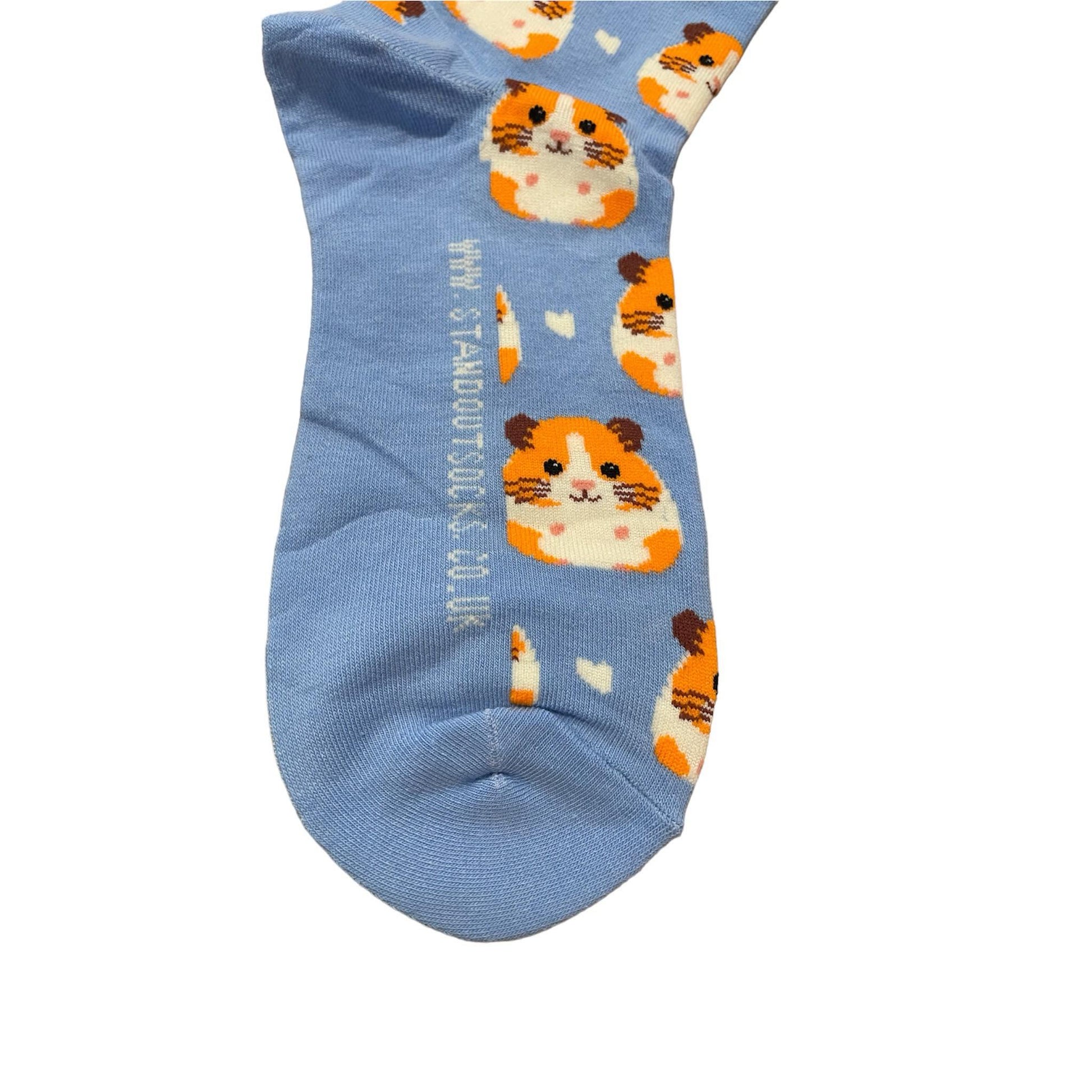 Light blue socks featuring a cute guinea pig pattern, created by Stand Out Socks. The socks display a playful design with orange and white hamster faces and small white hearts scattered throughout. The words "STAND OUT" are visible on the cuff and the company's website is printed on the sole.