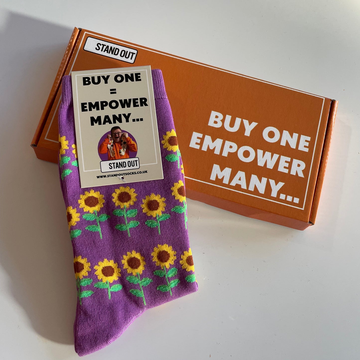 Purple socks with sunflower pattern on a white surface, featuring a cardboard box with text.