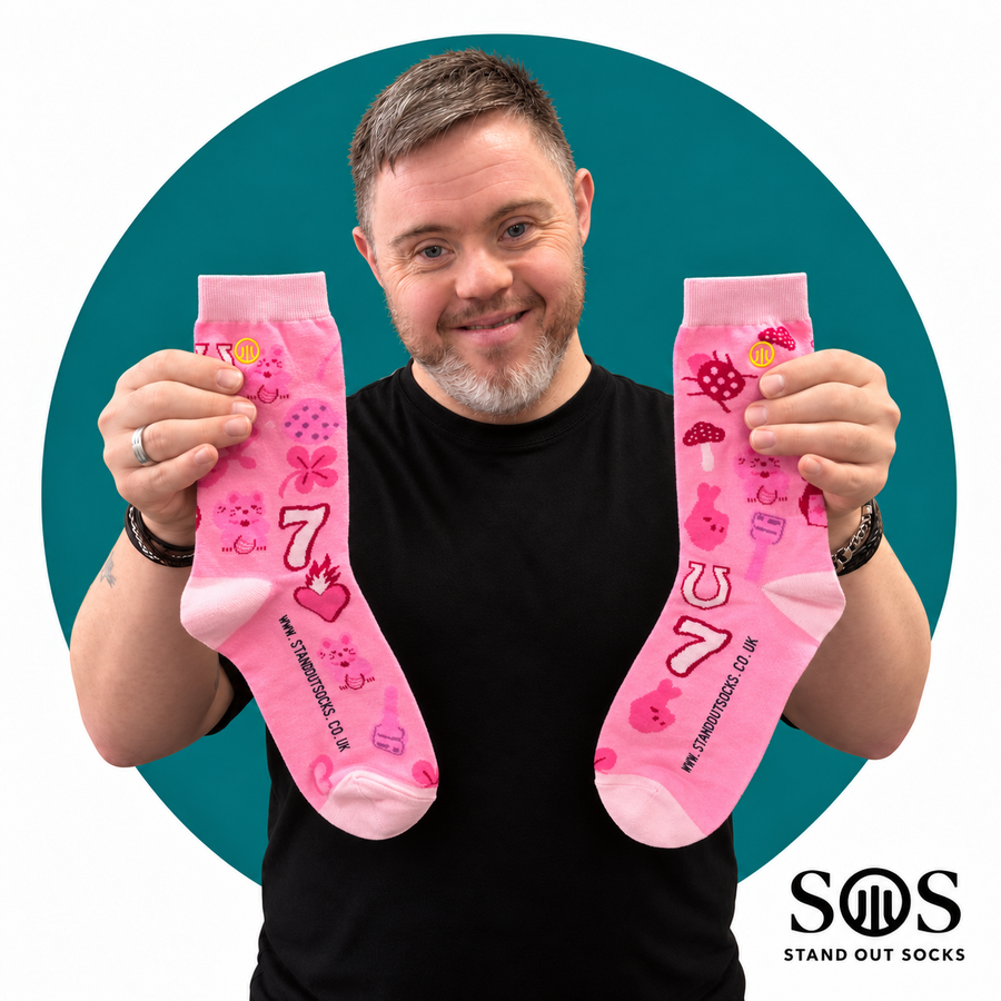 A smiling man holds up a pair of bright pink novelty socks with cartoon graphics and the number 7, set against a teal circular background with a white border. The socks feature pink bunnies, strawberries, and other playful motifs, with the brand 'Stand Out Socks' visible on the socks and as a logo in the bottom right corner.