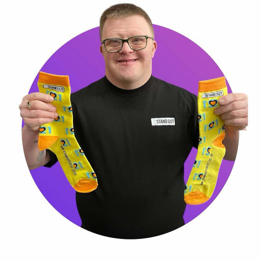 Cofounder of Stand Out Socks, Ross, Who has Down Syndrome, proudly holding up the Official World Down Syndrome Day Stand Out Socks which are yellow, blue and orange, feature a design that incorporates the number 21 mirrored to form three hearts at the centre, symbolising the triplication (trisomy) of the 21st chromosome, which causes Down syndrome.
