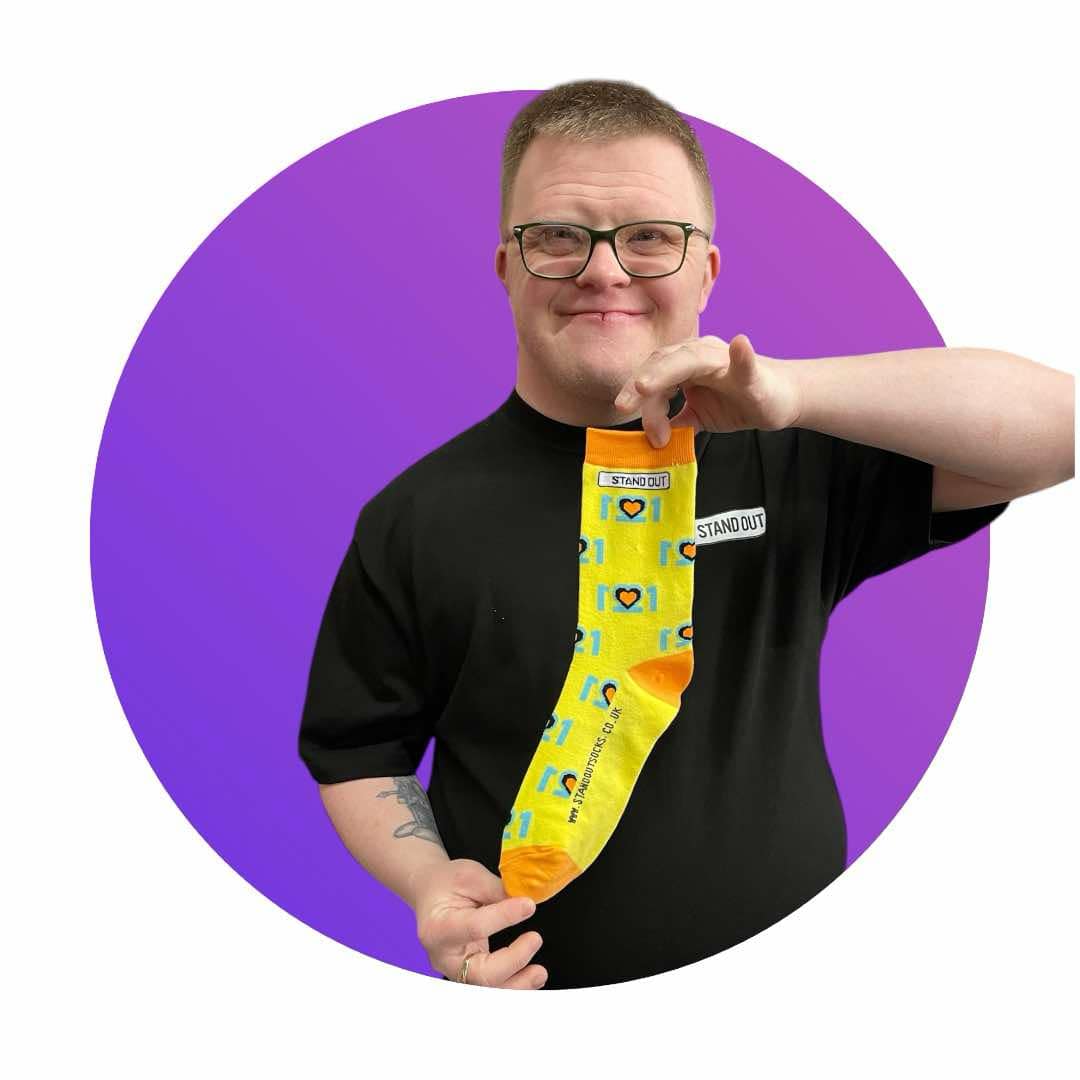 Cofounder of Stand Out Socks, Ross, Who has Down Syndrome, proudly holding up an Official World Down Syndrome Day Stand Out Sock which are yellow, blue and orange, feature a design that incorporates the number 21 mirrored to form three hearts at the centre, symbolising the triplication (trisomy) of the 21st chromosome, which causes Down syndrome.