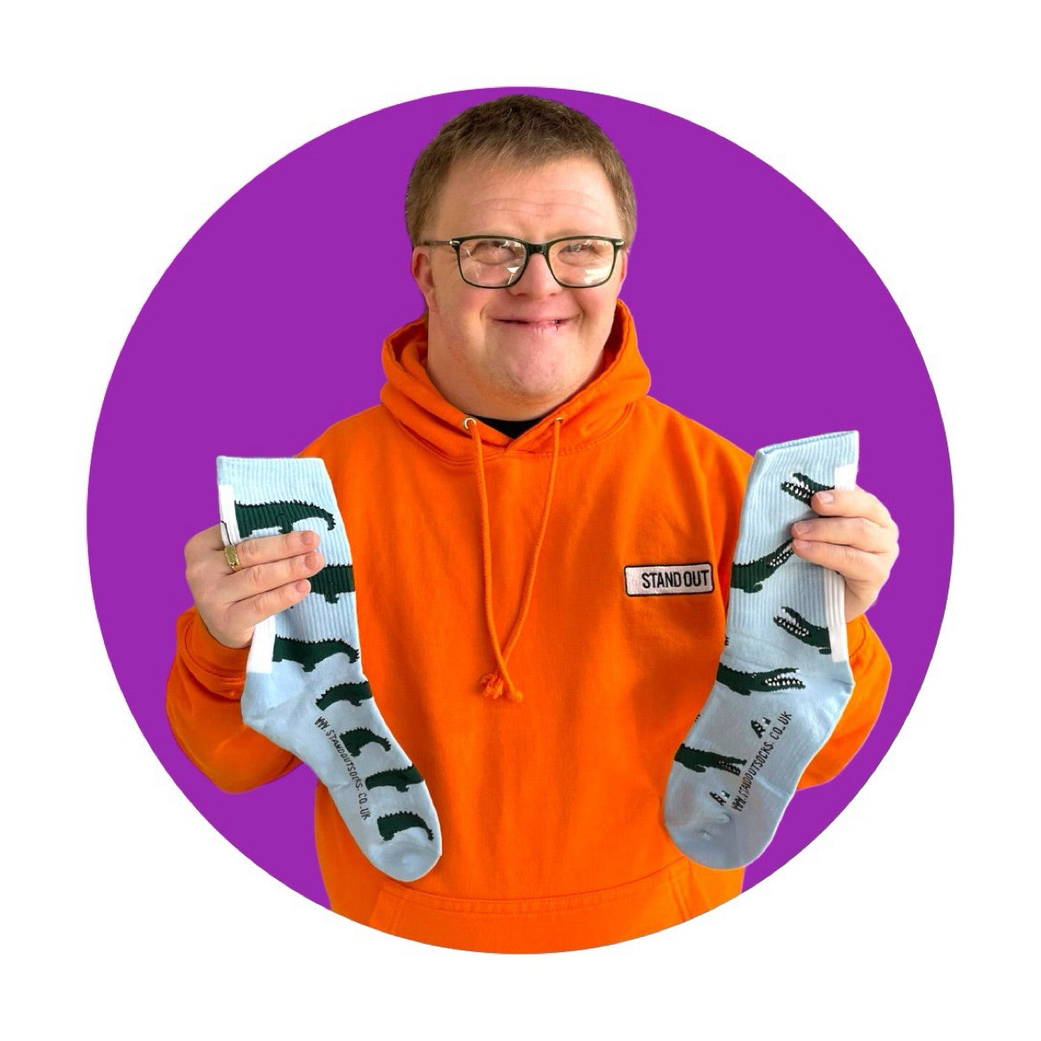 A man in an orange "Stand Out" hoodie proudly holding up a pair of colourful sports socks with unique design which is blue with green crocodiles. 