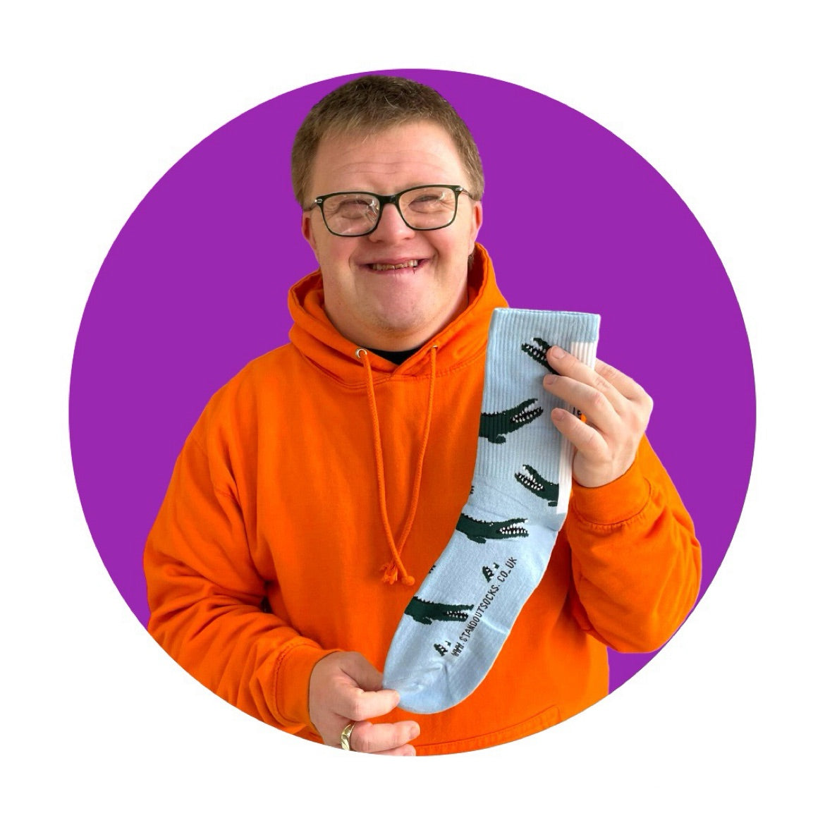 A man in an orange "Stand Out" hoodie proudly holding up a pair of colourful sports socks with unique design which is blue with green crocodiles. 
