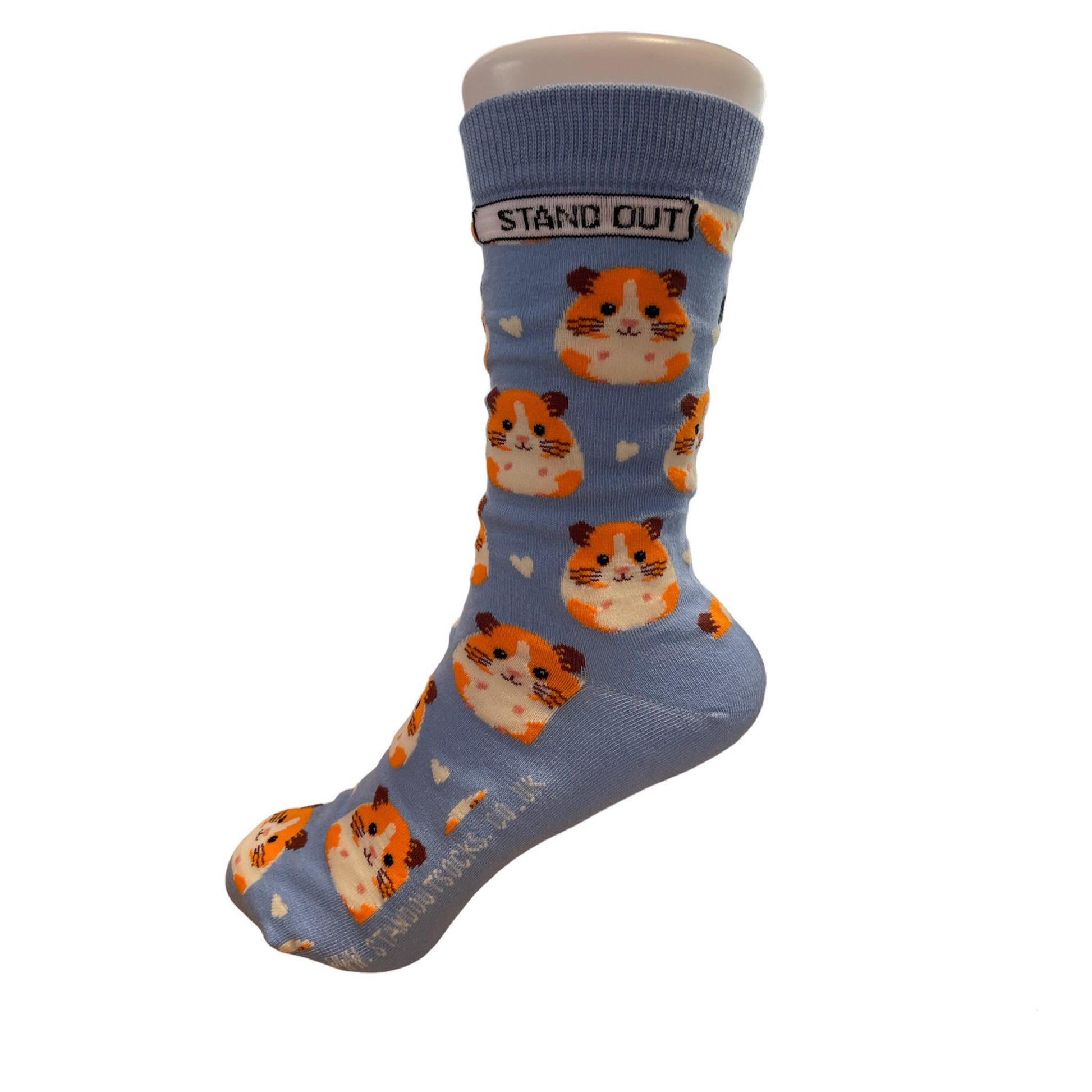 Light blue socks featuring a cute guinea pig pattern, created by Stand Out Socks. The socks display a playful design with orange and white hamster faces and small white hearts scattered throughout. The words "STAND OUT" are visible on the cuff and the company's website is printed on the sole.