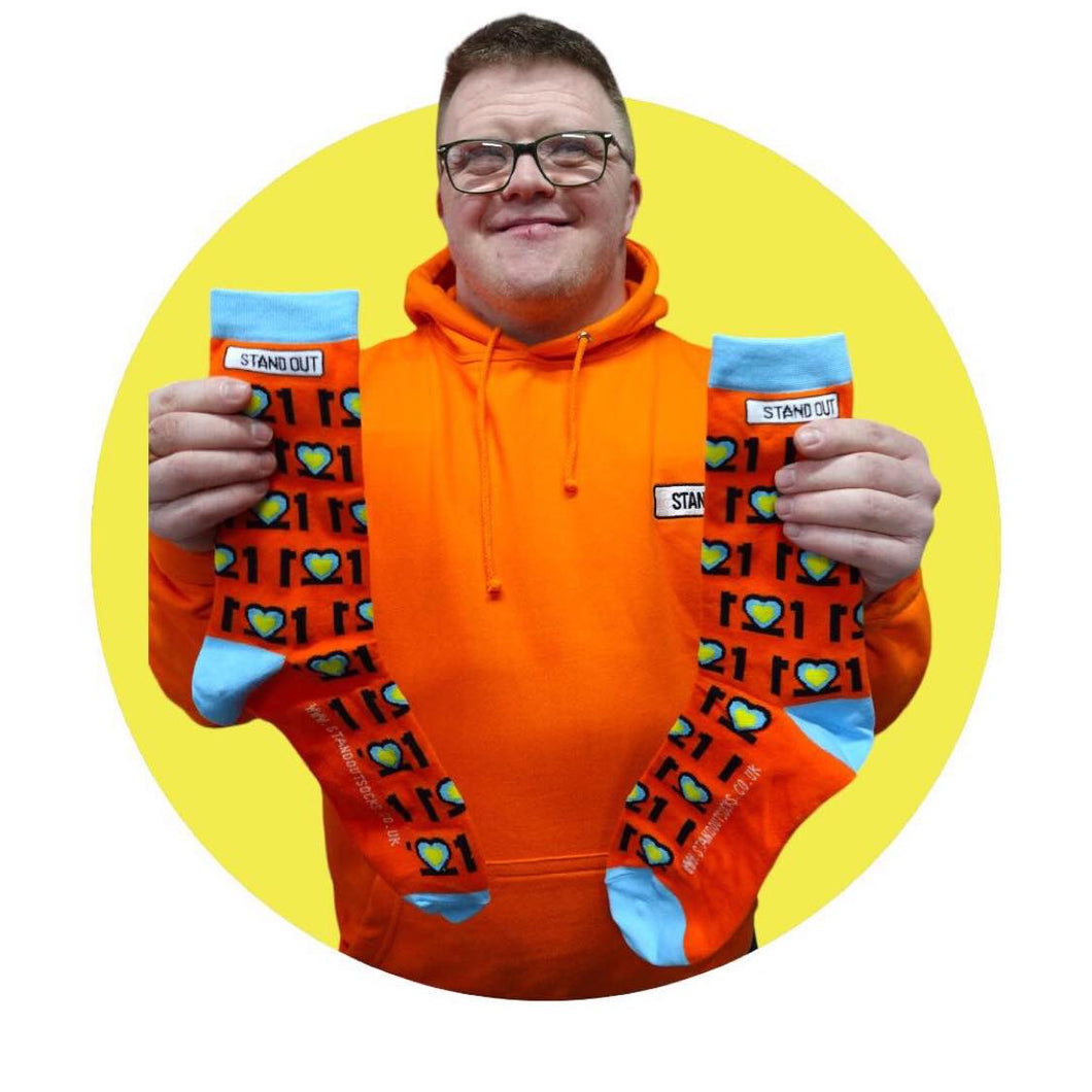 Man with Glasses on and orange jumper, Smiling and holding a pair of colourful Down Syndrome Awareness Socks