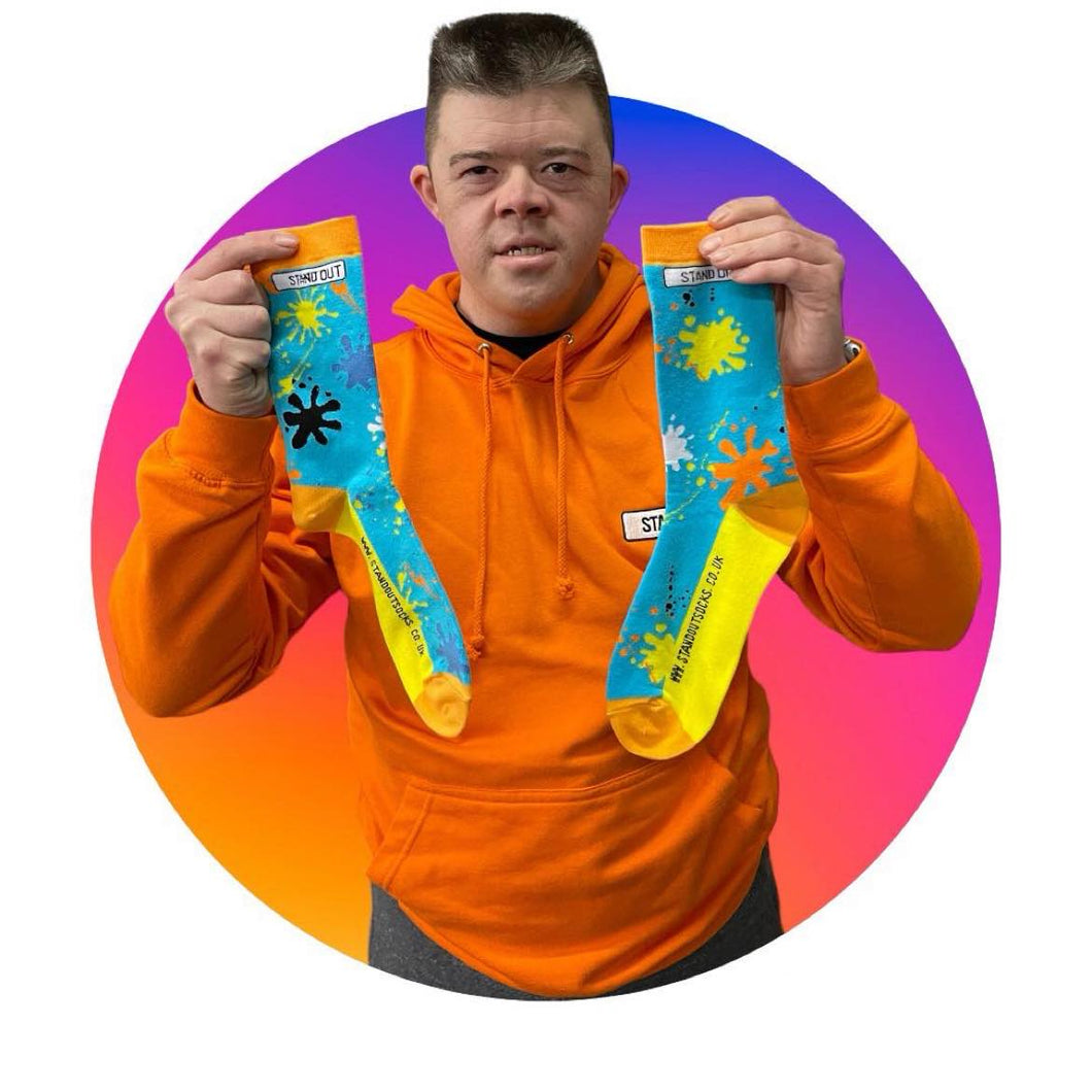 A man with Down Syndrome wearing an orange jumper on holding out a pair of funky colourful splat socks