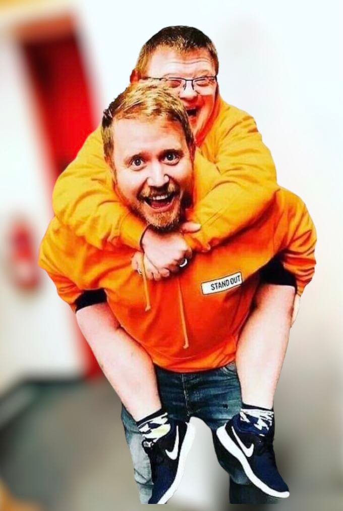 This image shows two men in bright orange hoodies with the "Stand Out" logo. The man in the foreground, with a big smile and light brown hair, is giving the man behind him, who has Down syndrome, a piggyback ride. Both are laughing joyfully, creating a warm and playful atmosphere. The man on the back is wearing glasses, and they both sport similar socks, contributing to the fun and lively feel of the image. The bright colours and their camaraderie reflect the spirit of inclusivity and positivity.
